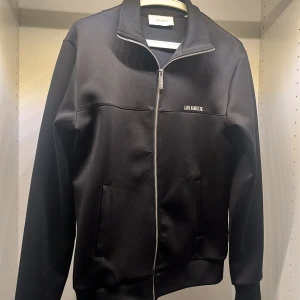 Svart track zip hoodie från Les Deux - Svart zip hoodie från Les Deux med stilren design och diskret logga på bröstet. Jackan har hel dragkedja framtill, ståkrage och två fickor. Materialet är mjukt och stretchigt, perfekt för en clean och stilig look.