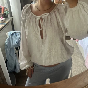 Vit blus med knyt och ballongärmar - Säljer en luftig vit blus med ballongärmar och snygg knytning vid halsen. Blusen har en lätt skrynklig struktur och är croppad i modellen, perfekt för en chill och trendig look. Passar dig som gillar boho-vibe och vill ha något bekvämt men ändå stilrent.