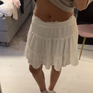 Vit volangkjol med resårmidja - Superfin vit kortkjol med flera volanger och resår i midjan. Kjolen har en luftig och flowy känsla, perfekt för varma dagar. Materialet känns lätt och bekvämt mot huden. Enkel att styla med både crop tops och stickade tröjor.