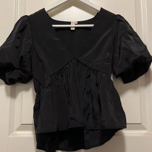 Svart omlottblus från Nelly - Supersnygg svart blus från Nelly med puffiga korta ärmar och omlottdetalj framtill. Blusen har en glansig finish och är tillverkad i ett silkesliknande material som ger en lyxig vibe. Perfekt för dig som gillar volym och coola detaljer.