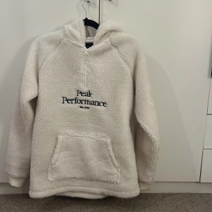 Vit fleecehoodie Peak Performance - Supermjuk och fluffig vit fleecehoodie från Peak Performance med broderad logga på bröstet. Tröjan har huva, magficka och half zip-dragkedja framtill. Perfekt för kalla dagar och chill vibes.