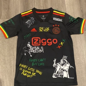 Ajax x Bob Marley svart fotbollströja - Unik svart Ajax fotbollströja från Adidas med Bob Marley-tema, Ziggo-logga och färgglada detaljer i rött, gult och grönt på ärmarna. Tryck med texter, illustrationer och musiknoter. Materialet är lätt och andas, perfekt för match eller träning.