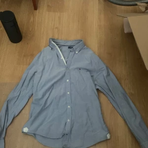 Blå skjorta från Tommy Hilfiger S - Klassisk ljusblå skjorta från Tommy Hilfiger i bomull. Skjortan har knappar framtill, lång ärm och en liten broderad logga på bröstet. Perfekt för en clean och stilren look med en snygg detalj ifall man kavlar upp ärmarna över en annan tröja. Storlek S