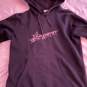 Saint Laurent hoodie - Säljer en svart hoodie från Saint Laurent med coolt tryck framtill i rosa och vit. Tröjan har huva med snörning och en stor magficka. Perfekt för dig som gillar streetstyle och vill ha något extra i garderoben. Pris kan diskuteras vid snabbaffär.