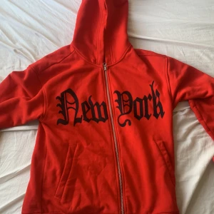 Röd hoodie med New York-print H&M XS - Röd hoodie från H&M i relaxed fit med svart New York-tryck fram och The New York City-tryck på ryggen. Tröjan har huva, dragkedja och fickor framtill. Materialet är en mix av bomull och polyester, perfekt för dig som gillar streetstyle.