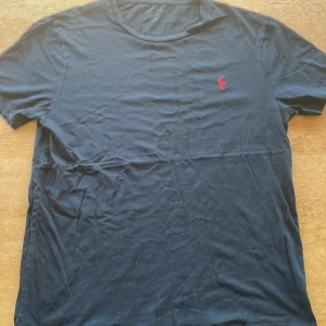 Svart t-shirt från Polo Ralph Lauren - Svart t-shirt från Polo Ralph Lauren med klassisk röd broderad logga på bröstet. Modellen är rak med rund hals och korta ärmar. Tillverkad i mjuk bomull som känns skön mot huden. Perfekt för dig som gillar stilrena och ikoniska plagg.