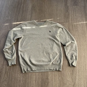 Grå stickad tröja från Polo Ralph Lauren - Snygg grå stickad tröja från Polo Ralph Lauren med klassisk rund hals och diskret logga broderad på bröstet. Tröjan har långärmade ribbade muddar och är perfekt för dig som gillar stilrena och tidlösa plagg. Passar dig som vill ha en chill och clean look.