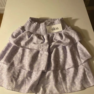 Ljuslila blommig volangkjol från Ellos - Superfin ljuslila kortkjol från Ellos med små vita blommor och flera volanger. Kjolen har bred, smockad midja som är stretchig och sitter skönt. Oanvänd. Öppen för bud!