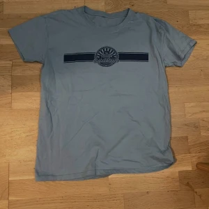 Blå t-shirt Sofialäkaren - Blå t-shirt från Sofialäkaren med tryckt logga och mörkblå rand över bröstet. Klassisk rund hals och korta ärmar. Perfekt för dig som gillar enkel och stilren design. Materialet känns mjukt och skönt mot huden.
