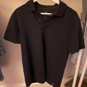 Blå pikétröja från Uniqlo M - Klassisk svart pikétröja från Uniqlo i storlek M. Tröjan har korta ärmar, krage och knäppning med två knappar framtill. Tillverkad i mjuk bomull som känns skön mot huden. Perfekt för en clean och stilren look.