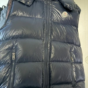 Mörkblå dunväst från Moncler - Snygg mörkblå dunväst från Moncler med glansig finish och ikonisk logga på bröstet. Västen har två praktiska fickor med dragkedja och detaljerade Moncler-zippar. Perfekt att slänga över en hoodie för extra värme under kyliga dagar. Denna västen är i bra skick förutom ett litet hål och borttagen qc lapp därav priset. Denna västen är köpt begagnad för ett tag sedan. Storlek 5 vilket motsvarar XL.                       Äkta solklart! Kontakta mig vid frågor. 