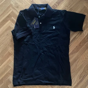 Svart Polo Ralph Lauren t-shirt M - Svart t-shirt från Polo Ralph Lauren med klassisk polokrage och två knappar. Slim fit-modell med vit broderad logga på bröstet. Tillverkad i mjuk bomull, perfekt för en clean och stilren look.