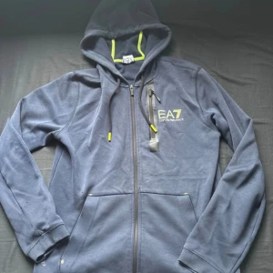 Marinblå EA7 hoodie med dragkedja - Säljer en marinblå hoodie från EA7 Emporio Armani med gul logga på bröstet och detaljer i gult. Tröjan har huva med snörning, hel dragkedja och en extra ficka med dragkedja på bröstet. Perfekt för chill eller träning.