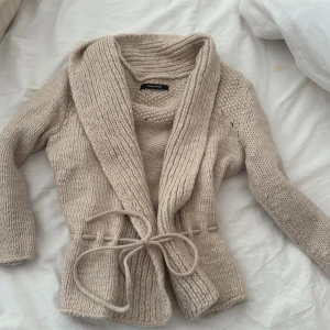 Beige stickad kofta från Samsøe Samsøe - Supermysig beige stickad kofta från Samsøe Samsøe med bred krage och knytband i midjan. Koftan har långärmade ärmar och grovstickad struktur upptill. Perfekt att slänga på sig när det är kyligt ute.