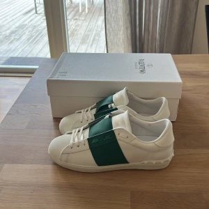 Valentino Garavani Open sneakers vit/grön - Snygga Valentino Garavani Open sneakers i vitt skinn med bred grön rem över mitten. Klassisk rund tå, vita snören och diskreta nitar på hälen. Perfekt för dig som vill sticka ut med exklusiva detaljer och stilren design.