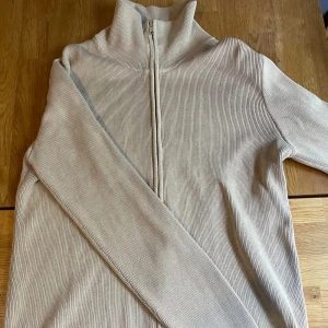 Beige ribbad kofta från Filippa K - Säljer en beige ribbad kofta från Filippa K i storlek XL. Tröjan har hel dragkedja framtill, hög krage och är tillverkad i 100% kammad bomull. Perfekt för dig som gillar stilrena och bekväma plagg med lång ärm och clean look. Har en liten fläck vid en av ärmarna - se sista bilden. 