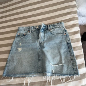 Blå jeanskjol från Stradivarius - Säljer en ljusblå jeanskjol från Stradivarius i storlek 34. Kjolen har rå kant med fransar, slitna detaljer framtill och klassiska fickor både fram och bak. Perfekt för dig som gillar en avslappnad och trendig stil.