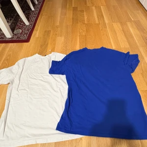 Blå och vit basic t-shirt - Två enkla t-shirts i klassisk modell, en i klarblått och en i vitt. Båda har rund hals och korta ärmar, perfekta för chill dagar eller när du vill ha en clean look. Materialet känns som mjuk bomull och passformen är normal.