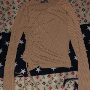 Beige ribbad långärmad topp Shein - Trendig beige långärmad topp från Shein med ribbad struktur och snörning på sidan som ger en snygg rynkad effekt. Croppad modell med rund halsringning, perfekt att styla med jeans eller kjol för en cool look.