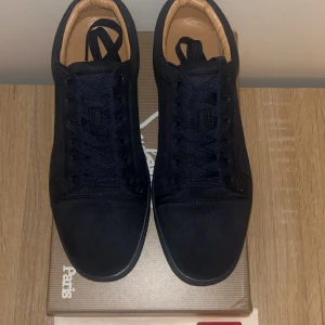 Svarta sneakers från Christian Louboutin - Snygga svarta sneakers från Christian Louboutin i mocka med klassisk röd sula. Skorna har rund tå, svarta snören och diskret broderad logga på hälen. Insidan är beige och de har en stilren, minimalistisk look som sticker ut med den ikoniska röda undersidan.