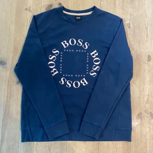 Mörkblå sweatshirt från Hugo Boss - Nyköps pris 1700kr,ca, Snygg mörkblå sweatshirt från Hugo Boss med stort BOSS-tryck i guld på bröstet. Tröjan har rund hals, långa ärmar och ribbade muddar. Perfekt för dig som gillar stilrena och trendiga plagg med premiumkänsla.