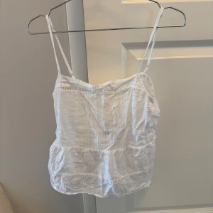 Vit linnetopp med knyt och volang - Supersöt vit linnetopp med tunna axelband, volangdetaljer och knyt i ryggen. Toppen har ett diskret rutigt mönster i tyget och öppen rygg. Perfekt för varma dagar och ger en luftig känsla.