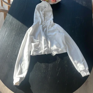 Vit croppad hoodie från H&M - Säljer en vit croppad hoodie från H&M i mjuk fleece. Tröjan har huva, dragkedja framtill och två fickor. Perfekt för dig som gillar en chill och trendig stil. Långärmad och med skön passform.