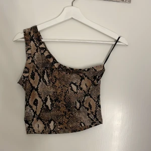 Oneshoulder topp med ormmönster - Snygg croppad topp med one shoulder och smal axelrem. Toppens ormmönster går i bruna, beige och svarta toner. Perfekt för dig som vill sticka ut med en trendig look.