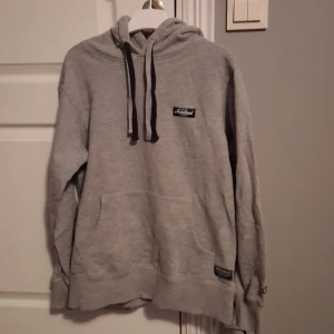 Grå hoodie från SQRTN Company - Säljer en grå hoodie från SQRTN Company med svart snörning och klassisk känguruficka. Tröjan har en broderad logga på bröstet och en patch nertill. Perfekt för chill dagar och enkel att matcha med jeans eller joggers.