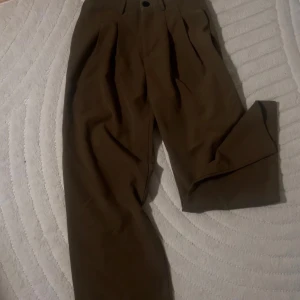 Bruna vida kostymbyxor - Säljer ett par bruna kostymbyxor med vida ben och hög midja. Byxorna har snygga veck framtill och stängs med knapp och dragkedja. Perfekta för en clean och stilren look.