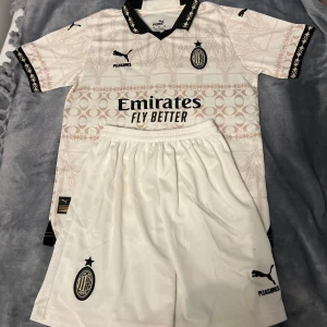 AC Milan fotbollströja och shorts Puma - AC Milan matchtröja och shorts i vitt med svarta detaljer. Tröjan har korta ärmar, svart krage och unikt mönster i beige. Puma-logga, klubbmärke och sponsortryck på bröstet. Shortsen är vita med klubbmärke och Puma-logga. Perfekt för fotbollsälskare!