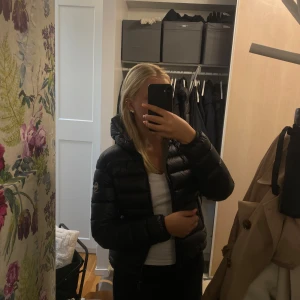 Svart dunjacka från Colmar - Säljer en svart, glansig dunjacka från Colmar med huva och dragkedja. Jackan har en puffig look, lång ärm och klassisk logga på ärmen. Perfekt för kalla dagar och riktigt snygg till streetwear-stil.