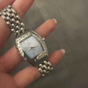 Silverfärgad klocka  - Stilren klocka köpt på en marknad i mallorca köpt för 500kr. Aldrig använt. silverfärgat armband i metall och rektangulärt ur med dekorativa detaljer runt boetten. Urtavlan är ljusblå och har tydliga siffror. Länkat armband med klassiskt spänne. Perfekt accessoar för att lyfta din outfit. Den måste ställas om. 