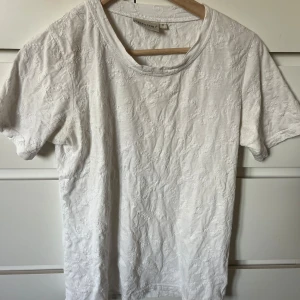 Vit blommig t-shirt från Cappuccini - Vit t-shirt från Cappuccini i storlek M med korta ärmar och rund hals. T-shirten har ett diskret blommigt broderat mönster över hela tyget som ger en unik och fräsch vibe. Materialet känns mjukt och är troligen bomull, perfekt för dig som gillar stilrena detaljer.