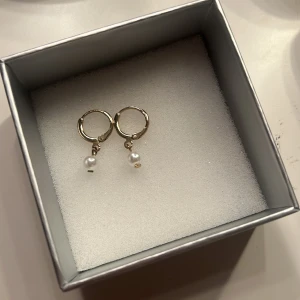 Guldiga örhängen med pärlor - Snygga små guldiga hoops med hängande vita pärlor. Örhängena är runda och har en enkel design som ändå sticker ut tack vare pärldetaljen.