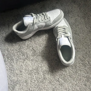 Nike Dunk Low i grått och vitt - Nike Dunk Low sneakers i stilren grå och vit färg. Klassisk siluett med rund tå, perforerad tåbox och snörning. Ovandel i skinn med ikonisk Swoosh på sidorna. Perfekta för dig som gillar clean och tidlös streetstyle.
