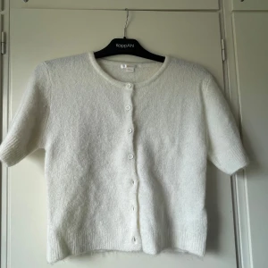 Vit stickad kofta från Gina Tricot - Supermjuk och fluffig vit stickad kofta från Gina Tricot med kort ärm och rund hals. Koftan har knappar framtill och kort ärm! Inte stickig, använd en gång❣️❣️