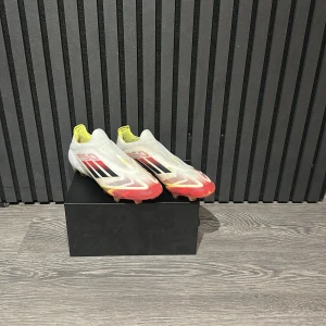 Adidas F50 elite fotbollsskor i vitt och rött - Säljer ett par Adidas F50 fotbollsskor i vitt med röda och gula detaljer samt svarta ränder på sidorna. Skorna har en strömlinjeformad design utan snörning och är tillverkade i syntetmaterial för lätt känsla på planen. Sulan har gula dobbar för bra grepp.