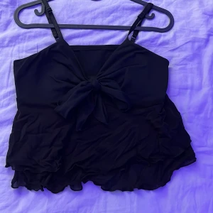 Svart volangig linnetopp med rosett - Supersöt svart linnetopp med volanger och stor rosett framtill. Toppen har smala axelband och är tillverkad i ett lätt, flowy material som ger en luftig känsla. 