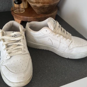 Vita Nike Air Jordan 1 Lows - Snygga helvita Nike Air Jordan 1 Lows med klassisk design och perforerad tå. Skorna har snörning och är tillverkade i skinn med diskret Nike-logga på sidan. Perfekta för dig som gillar stilrena sneakers med ikonisk känsla.