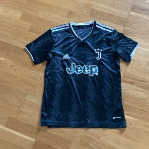 Juventuströja Adidas xs/s - Juventus svart fotbollströja från Adidas med vita detaljer och V-ringad krage. Tröjan har korta ärmar, subtilt mönster och klassiska Adidas-ränder på axlarna. Jeep-logga och Juventus-emblem med tre stjärnor på bröstet. Tillverkad i lätt polyester.