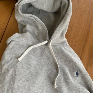 Grå hoodie från Polo Ralph Lauren - Säljer en grå hoodie från Polo Ralph Lauren med klassisk svart logga på bröstet. Tröjan har huva med vit snörning, känguruficka och ribbade muddar. Perfekt för chill dagar och enkel att matcha med jeans eller joggers. Hoodien har ett par små fett fläckar som synns knapt ovanför fickan.