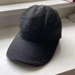 Svart keps Fendi  - Exklusiv svart keps från Fendi med diskret FF-monogram i tyget.  har justerbar rem baktill med Fendi-logga. Snygg och stilren design som lyfter vilken outfit som helst. Helt ny och oandvänd.