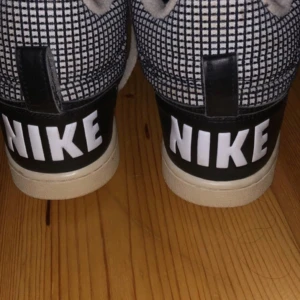 Nike svartvita rutiga sneakers high - Snygga Nike sneakers med svart och vitt rutigt mönster på sidorna, svarta skinnpaneler och stora vita NIKE-bokstäver på hälen. Skorna har högt skaft, vita snören och mjuk vadderad insida. Perfekta för dig som gillar streetstyle och vill sticka ut.