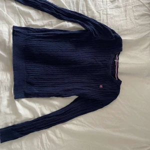 Mörkblå stickad tröja Gina Tricot - Mörkblå kabelstickad tröja från Gina Tricot i storlek M. Tröjan har rund halsringning, långa ärmar och en liten rosa broderad detalj på bröstet. Ribbstickade muddar vid ärmslut och nederkant. Perfekt för lager-på-lager under kyliga dagar.