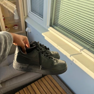 Svarta Nike Air Force 1 sneakers - Säljer ett par helsvarta Nike Air Force 1 sneakers med klassisk design. Skorna har snörning, perforerad tå och är tillverkade i skinn med en platt sula. Perfekta för dig som gillar en clean och tidlös look.