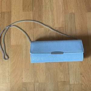 Glittrig ljusblå kuvertväska Anna Field - Snygg ljusblå kuvertväska från Anna Field med glittrig yta och silverfärgad kedjerem. Väskan har ett stilrent metallspänne och är perfekt för att lyfta din outfit. Insidan har ett litet fack för kort eller småsaker.