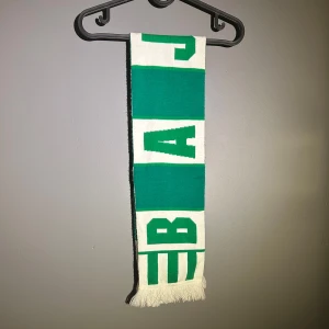 Hammarby halsduk - Snygg halsduk i grönt och vitt med tydlig text och blockmönster. Perfekt för dig som vill sticka ut på läktaren eller bara gillar sportig stil. Halsduken har fransar i ändarna och är tillverkad i ett mjukt stickat material. 