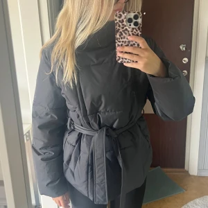 Grå pufferjacka med bälte - Säljer en grå pufferjacka med bälte i midjan och hög krage. Jackan har en clean look, långa ärmar och är quiltad för extra värme. Perfekt för kalla dagar och ger en snygg siluett. Passformen är normal och jackan är riktigt skön att bära. Köpt från Vero Moda för 800!