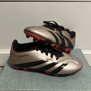 Adidas Predator fotbollsskor guld/svart - Snygga Adidas Predator fotbollsskor i metallic guld med svarta detaljer och röda dobbar. Skorna har klassisk snörning och de ikoniska tre svarta ränderna på sidan. använt i typ tre dagar, säljer för att de inte passade. Köptes för 1500 säljs för 700 skriv om pris och mer ni undrar över.
